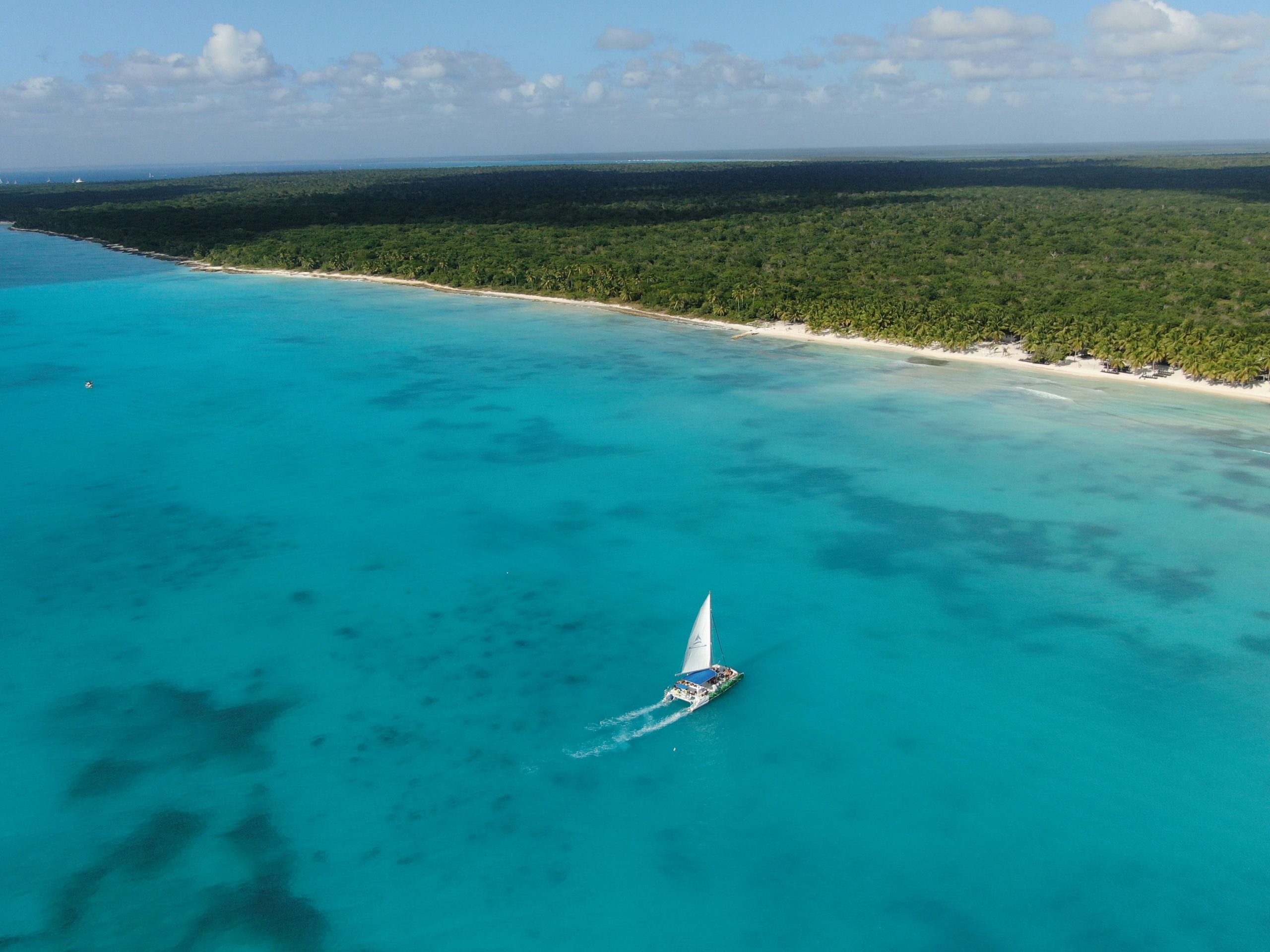 A sailboat glides along a pristine turquoise shoreline under a sunny sky.